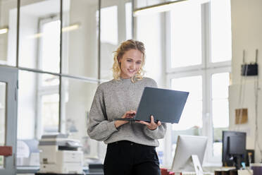 Lächelnde junge Frau, die im Büro steht und einen Laptop benutzt - MMIF00329