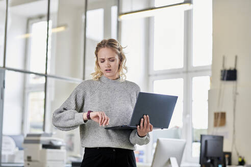 Young woman standing in office with laptop checking the time - MMIF00330