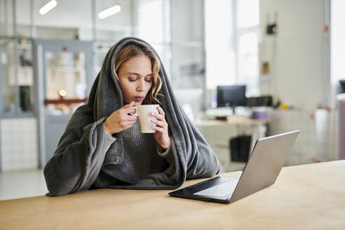 Junge Frau in gemütlicher Loungewear sitzt am Schreibtisch im Büro und trinkt aus einer Tasse - MMIF00334