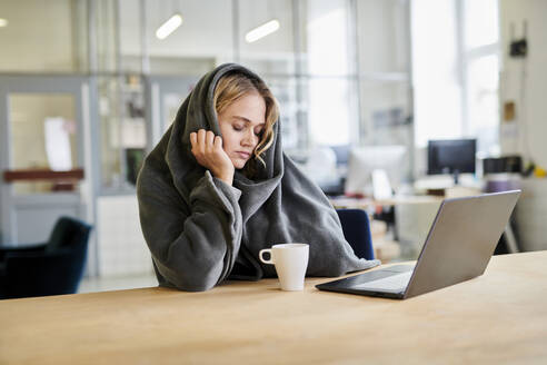 Exhausted young woman in cozy loungewear sitting at desk in office with laptop - MMIF00335