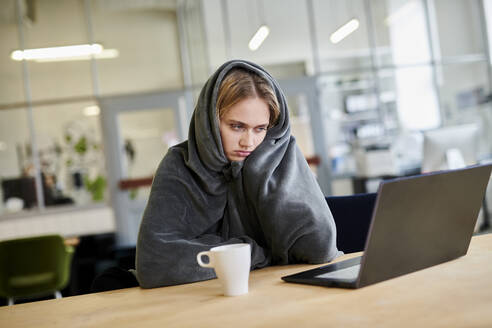 Frustrierte junge Frau in gemütlicher Loungemode sitzt am Schreibtisch im Büro mit Laptop - MMIF00336