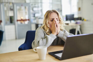 Frustrierte junge Frau sitzt am Schreibtisch im Büro mit Laptop - MMIF00339