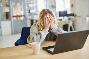 Verzweifelte junge Frau sitzt am Schreibtisch im Büro mit Laptop - MMIF00340