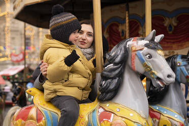 Frau stützt einen Jungen, der auf einem Karussellpferd auf dem Weihnachtsmarkt sitzt - SSGF00961
