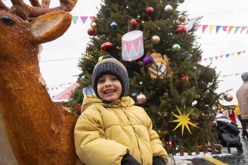 Fröhlicher Junge neben Hirschdekoration auf dem Weihnachtsmarkt - SSGF00963
