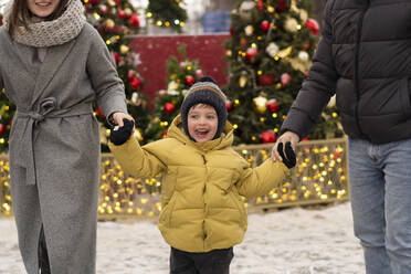 Lächelnder Junge hält die Hände seiner Eltern auf dem Weihnachtsmarkt - SSGF00967