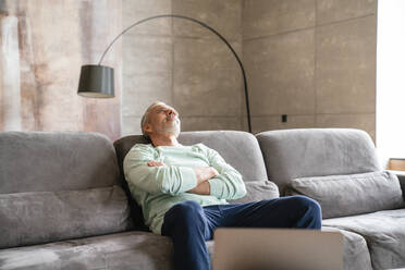 Thoughtful mature man with arms crossed sitting on sofa at home - VPIF06072