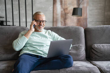 Smiling businessman holding laptop talking on mobile phone at home - VPIF06078