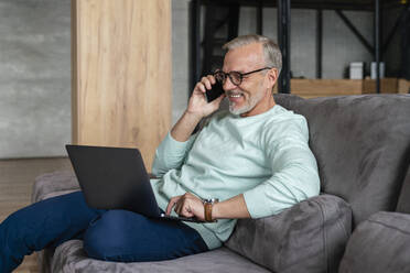 Happy businessman holding laptop talking on mobile phone at home - VPIF06079