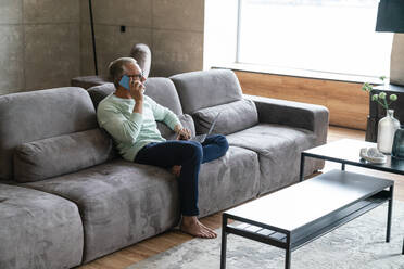 Businessman holding laptop talking on mobile phone at home - VPIF06080