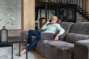 Businessman talking on smart phone sitting with laptop on sofa at home - VPIF06081