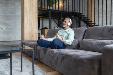 Man listening music through wireless headphones practicing yoga on sofa - VPIF06097