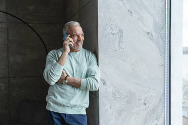 Smiling man talking on phone standing by window at home - VPIF06107