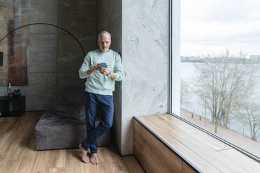 Älterer Mann mit Smartphone am Fenster stehend zu Hause - VPIF06108