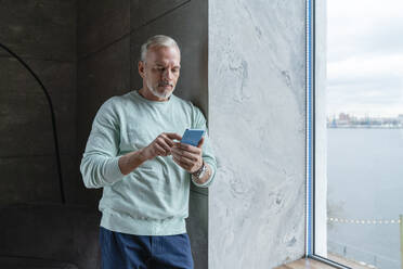 Mature man using mobile phone standing by window at home - VPIF06109