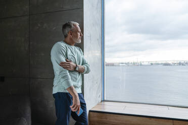 Nachdenklicher Mann mit Handy in der Hand, der durch das Fenster seines Hauses schaut - VPIF06110