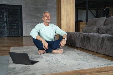 Mann mit geschlossenen Augen macht Yoga am Laptop auf dem Teppich zu Hause - VPIF06133