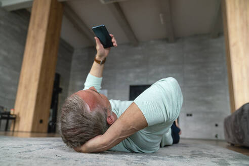 Älterer Mann mit Hand hinter dem Kopf, der ein Smartphone benutzt und zu Hause auf dem Teppich liegt - VPIF06144