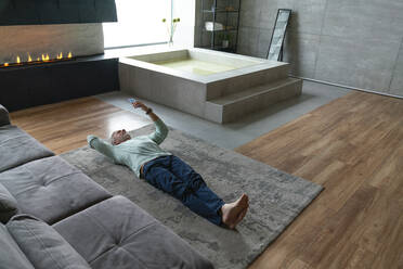 Mann mit Mobiltelefon auf Teppich im Wohnzimmer liegend - VPIF06145