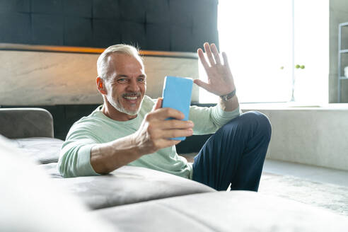 Lächelnder Mann bei einem Videogespräch, der mit seinem Smartphone im Wohnzimmer winkt - VPIF06149