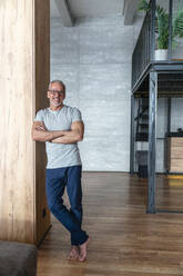 Smiling man with arms crossed leaning on wooden column at home - VPIF06179