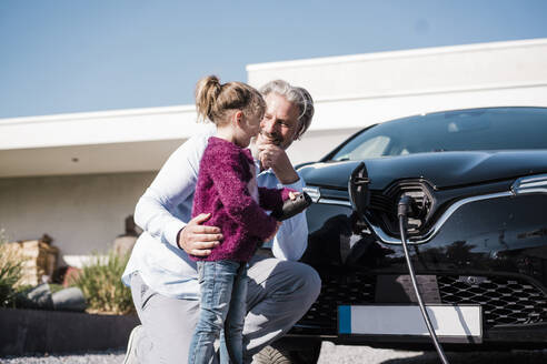 Glückliches Mädchen, das einen Stromstecker hält und neben seinem Vater vor einem Auto steht, an einem sonnigen Tag - MOEF04069