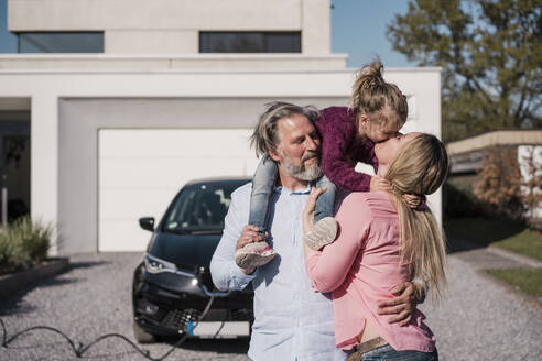 Frau küsst Tochter, die auf der Schulter eines Mannes vor einem Elektroauto sitzt - MOEF04076