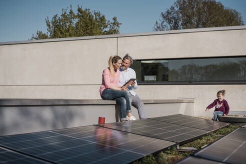 Frau mit Tablet-PC sitzt neben Mann mit Tochter, die an einem sonnigen Tag an einem Sonnenkollektor spielt - MOEF04093