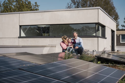 Glückliches Mädchen mit Eltern, die an einem Sonnenkollektor auf dem Dach sitzen - MOEF04095