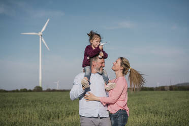 Lächelnder Mann, der seine Tochter auf der Schulter trägt und ein Modell einer Windturbine hält, die neben einer Frau auf einem Bauernhof steht - MOEF04098