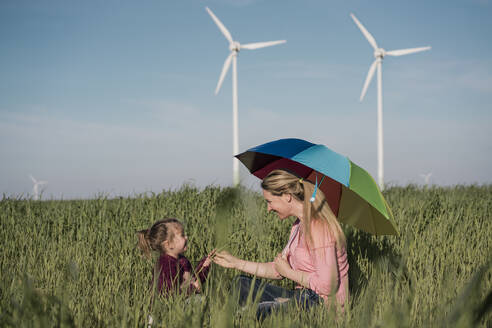 Smiling girl and mother sitting with colorful umbrella in in field - MOEF04116