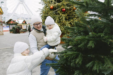 Vater und Töchter hängen Weihnachtsschmuck an den Baum auf dem Markt - SIF00124