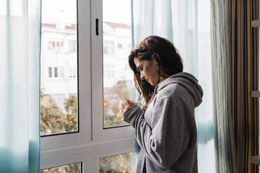 Mature woman with mug of tea standing by window at home - DCRF01182