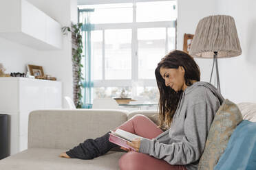 Mature woman reading book sitting on sofa in living room at home - DCRF01190