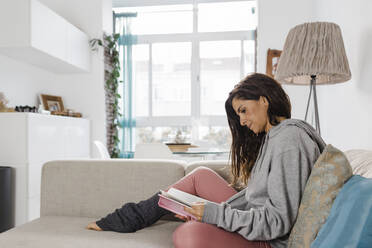 Reife Frau, die ein Buch liest, sitzt auf dem Sofa im Wohnzimmer zu Hause - DCRF01190