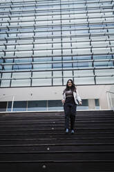 Lächelnde Geschäftsfrau, die sich auf einer Treppe vor einem modernen Gebäude nach unten bewegt - DCRF01206
