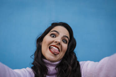 Playful of happy woman sticking out tongue taking selfie in front of blue wall - DCRF01211