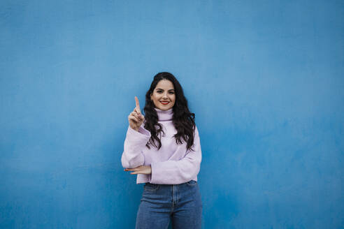 Smiling young beautiful woman showing number one with index finger in front of blue wall - DCRF01221