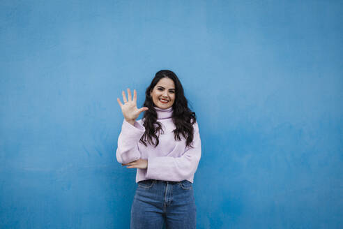 Smiling young beautiful woman showing number 5 in front of blue wall - DCRF01225