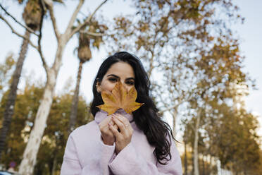 Young woman covering face with autumn leaf at park - DCRF01234