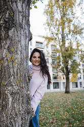 Smiling young beautiful woman enjoying autumn standing behind tree trunk at park - DCRF01237