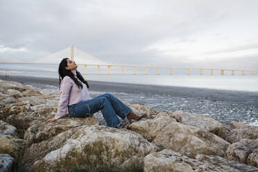 Young woman sitting on rocks relaxing at beach by river - DCRF01239
