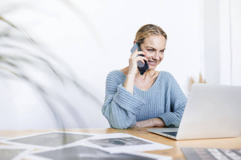 Lächelnde Geschäftsfrau, die mit dem Handy telefoniert und zu Hause am Laptop sitzt - RFTF00202