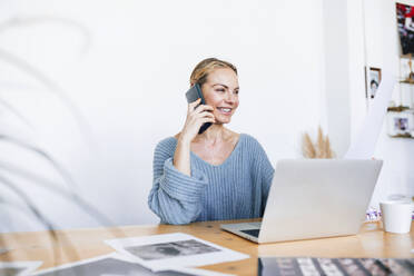 Happy businesswoman talking on mobile phone sitting with laptop at home - RFTF00203