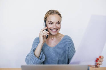 Smiling businesswoman holding document talking on mobile phone sitting with laptop at home - RFTF00206