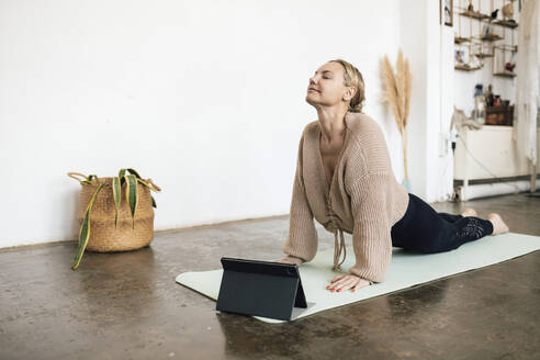 Frau übt Kobra-Pose auf Übungsmatte vor Tablet-PC zu Hause - RFTF00213