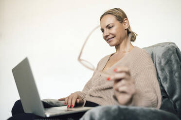 Lächelnde Geschäftsfrau mit Brille und Laptop auf einem Sessel zu Hause - RFTF00216