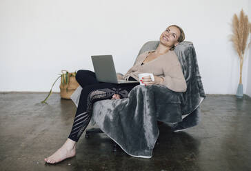 Lächelnde Frau mit Kaffeetasse und Laptop auf einem Sessel zu Hause - RFTF00217