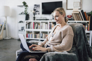 Woman using laptop sitting on armchair at home - RFTF00219