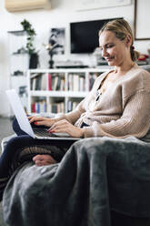 Smiling woman using laptop sitting on armchair at home - RFTF00220